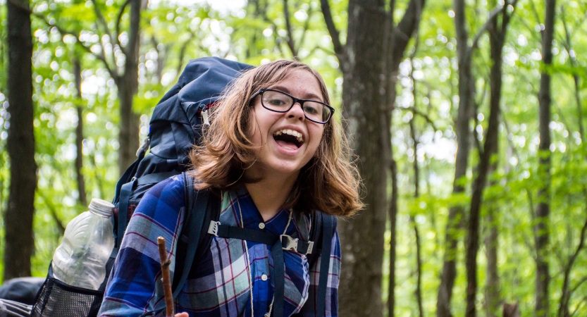 A person wearing a backpack smiles for the photo amidst a green wooded area 
